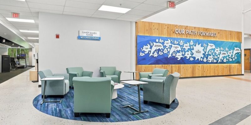 Brown Family YMCA wellness lounge with green seating, small tables and a donor recognition wall.